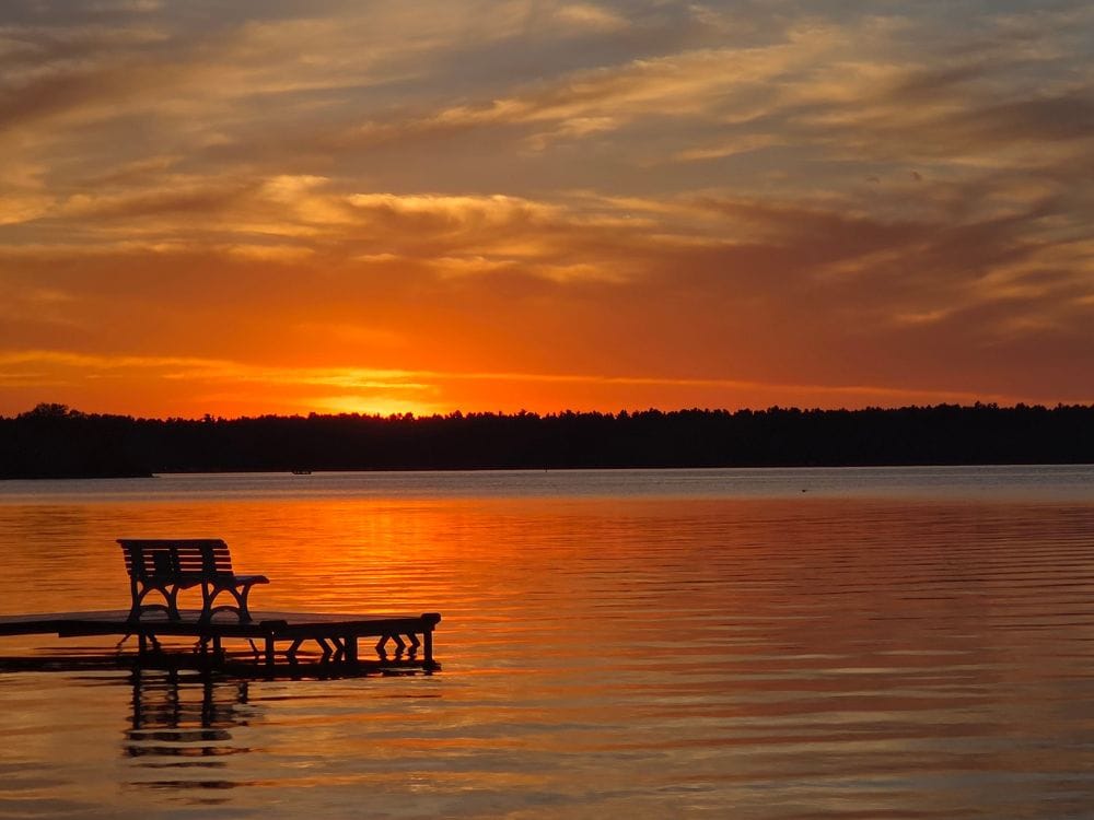 A quiet bench sits at the end of a dock overlooking a calm lake at sunset with warm light reflecting across the water. The image symbolizes reflection, personal values, and the thoughtful decision making involved when choosing between cremation vs. burial and considering what matters most in end of life planning.
